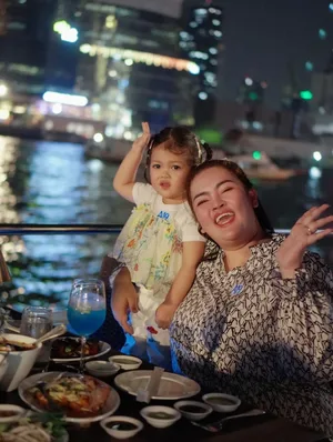 ผู้หญิงและเด็กเล็กโพสท่าข้างโต๊ะอาหารบนเรือ มีแม่น้ำและแสงไฟอาคารเป็นฉากหลัง