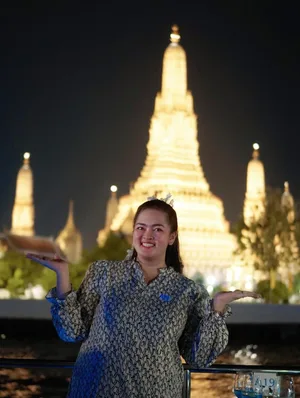 ผู้หญิงยิ้มและยกมือสองข้างบนเรือ มีพระปรางค์วัดอรุณประดับไฟเป็นฉากหลัง