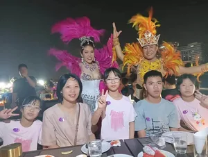 กลุ่มผู้โดยสารถ่ายภาพที่โต๊ะอาหารร่วมกับนักแสดงสองคนในชุดขนนกสีชมพูและสีส้ม