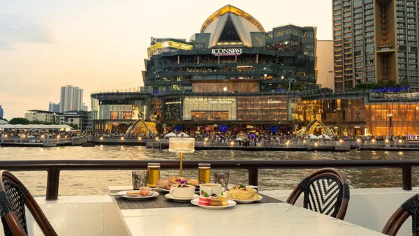 อาหารและขนมบนโต๊ะริมเรือ The Luxury White รอบ Twilight มองเห็น ICONSIAM ที่เปิดไฟในช่วงเย็น