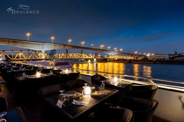 โต๊ะริมดาดฟ้า The Opulence Cruise พร้อมโคมไฟสีขาว มองเห็นสะพานและแสงสะท้อนสีทองบนแม่น้ำช่วงฟ้าสีน้ำเงิน