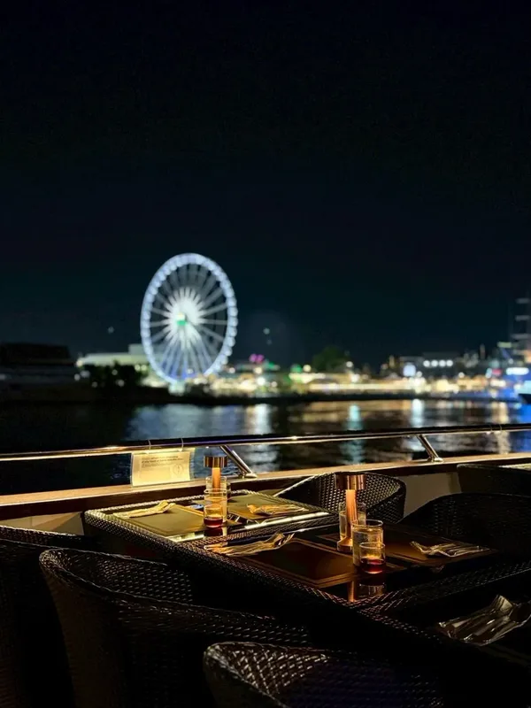 โต๊ะริมเรือ The Opulence Cruise พร้อมโคมไฟและแก้วเครื่องดื่ม มีชิงช้าสวรรค์เอเชียทีคเป็นฉากหลังยามค่ำคืน