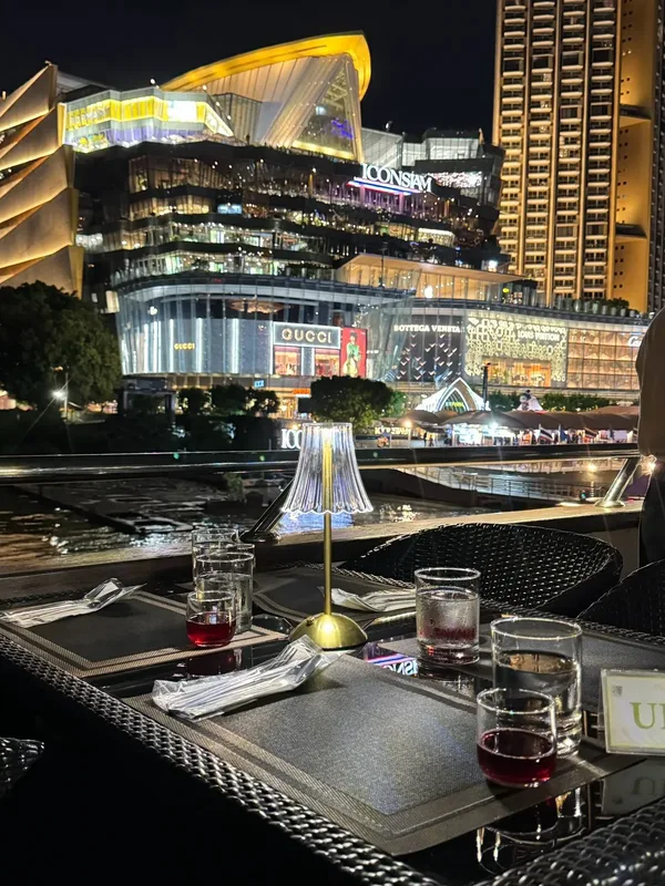 โต๊ะริม The Opulence Cruise รอบ Dinner มีโคมไฟทรงจีบและแก้วเครื่องดื่ม โดยมีไอคอนสยามอยู่ด้านหลัง