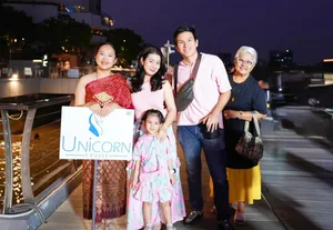 กลุ่มผู้โดยสารผู้ใหญ่สามคนและเด็กถ่ายภาพกับพนักงานชุดไทยที่ถือป้าย Unicorn Cruise บริเวณทางเดินริมน้ำ
