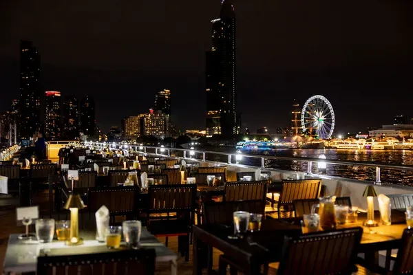 โต๊ะอาหารริมดาดฟ้าเรือ Viva Alangka Cruise ประดับโคมไฟสีทอง มองเห็นชิงช้าสวรรค์เอเชียทีคและแสงไฟริมแม่น้ำเจ้าพระยายามค่ำคืน