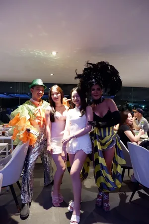 ผู้โดยสารหญิงสองคนถ่ายภาพกับนักแสดงในชุดสีสันสดใสและเครื่องศีรษะขนนกสีดำในห้องอาหารเรือ Viva Alangka Cruise