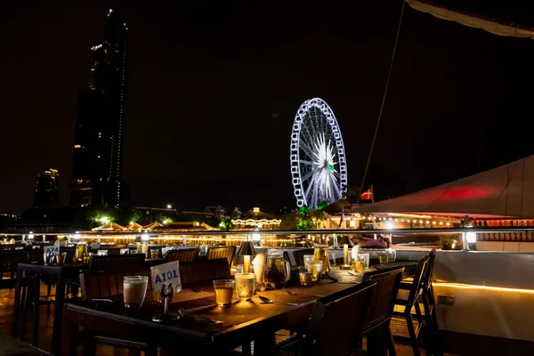 โต๊ะอาหารริมดาดฟ้าเรือ Viva Alangka Cruise จัดแก้วน้ำและโคมไฟโทนอุ่น พร้อมวิวชิงช้าสวรรค์เอเชียทีคที่ส่องสว่างยามค่ำคืน