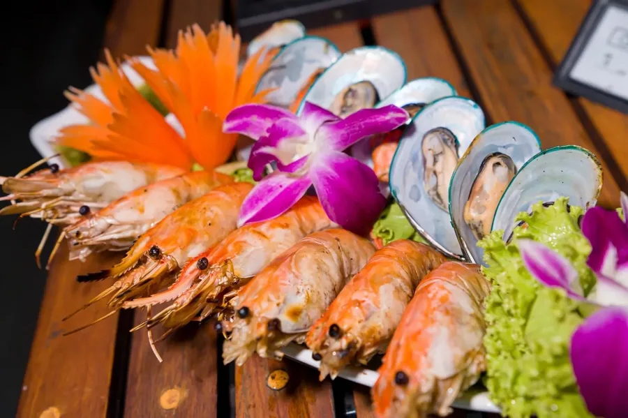 กุ้งและหอยแมลงภู่จัดเรียงคู่กันบนจาน ตกแต่งด้วยกล้วยไม้สีม่วง เพิ่มสีสันให้มื้อซีฟู้ดบนเรือ